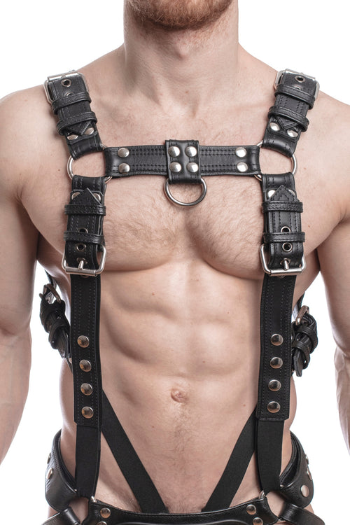 Model wearing a black leather combat harness and connector with stainless steel hardware, connected to a jock and a cockring. Front view.