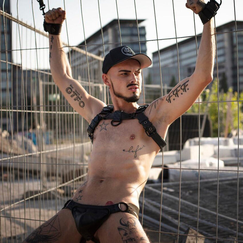 Model wearing black leather combat bulldog chest harness and leather jockstrap chained to a fence
