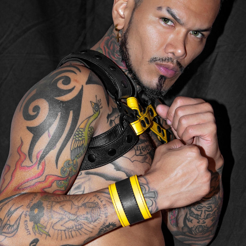 Heavily tattooed male model wearing a yellow and black leather striped wristband with matching coloured chest harness