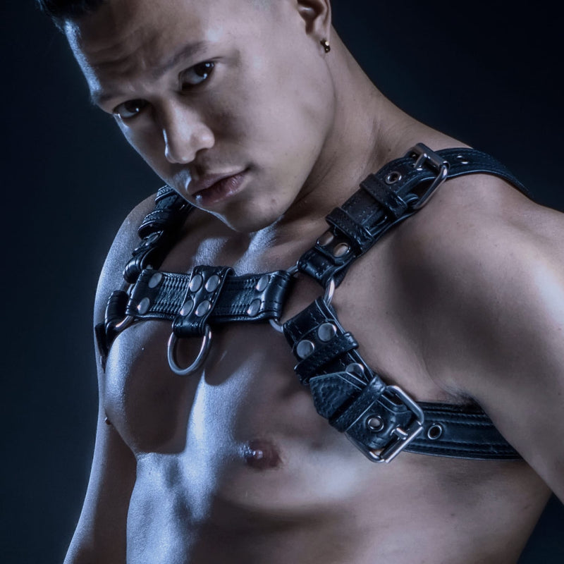 Model wearing a black leather chest harness with detailed stitching and stainless steel hardware.
