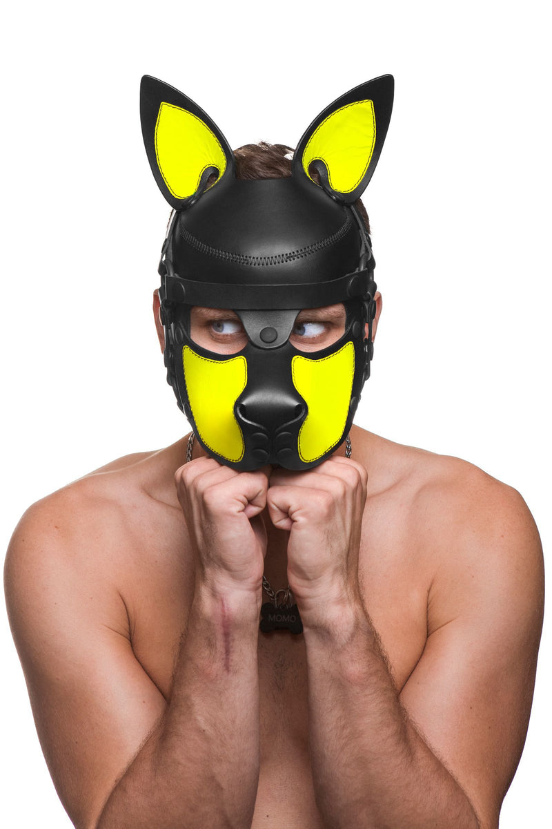 Waggish model in a cute black and neon yellow leather pup hood with his chin resting on his hands is looking off to the left.