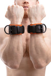 Male model displaying a pair of black and orange leather wrist restraints with black metal rivets, buckles, and D-rings.
