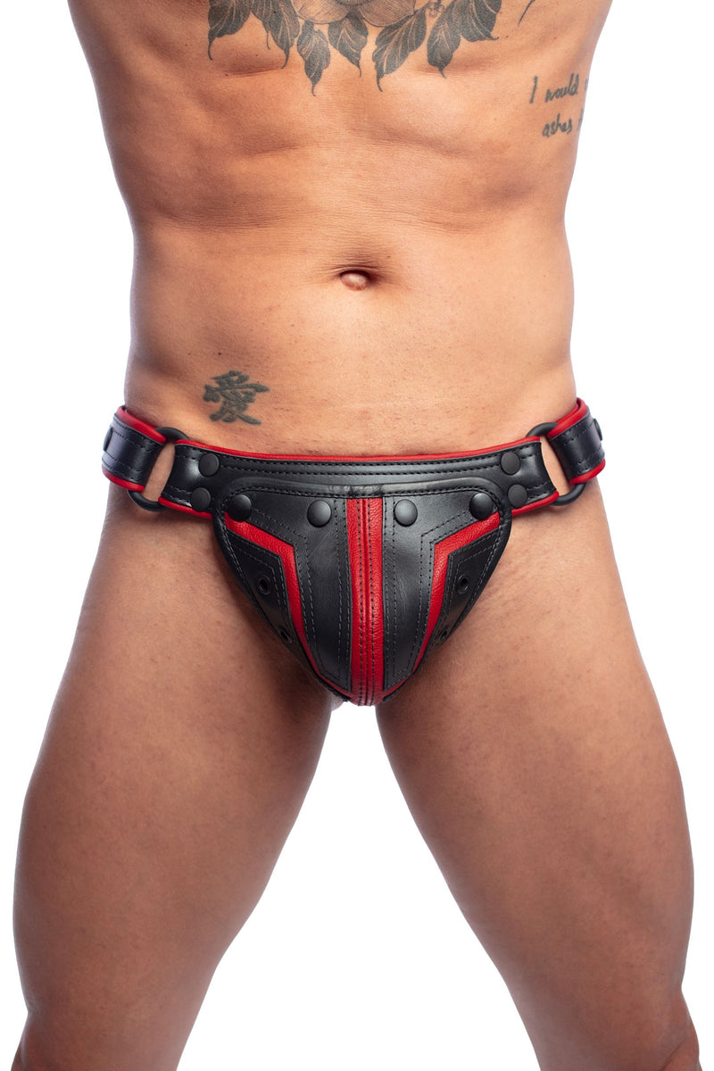 A toned model wearing a black and red leather Combat thong with matte black rivets, snaps, and O-rings, front facing view.