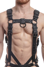 Model wearing a black leather combat harness and connector with black metal hardware, connected to a jock and a cockring. Front view.