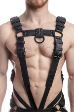 Model wearing a black leather combat harness and connector with black metal hardware, connected to a cockring. Front view.