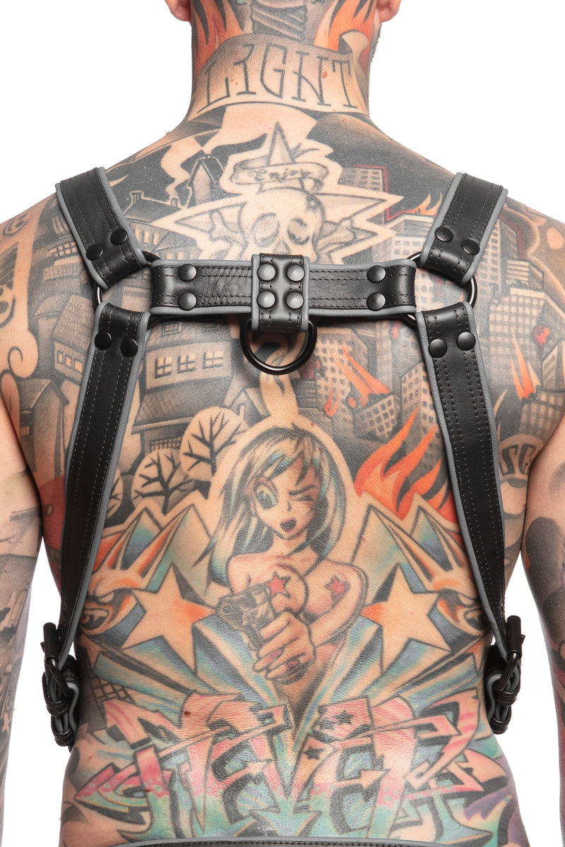 Model wearing a black and grey leather combat harness and connector with black metal hardware. Back view.