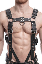 Model wearing a black leather combat harness and connector with stainless steel hardware, connected to a jock and a cockring. Front view.