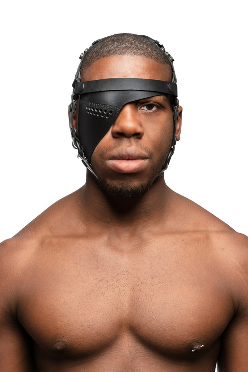Model wearing black leather head harness and right eye patch with black metal hardware. Front view.