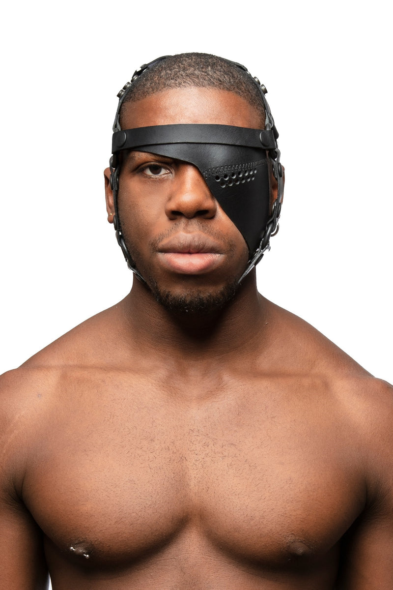 Model wearing black leather head harness and left eye patch with black metal hardware. Front view.