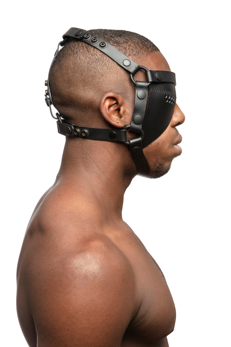 Model wearing black leather head harness and right eye patch with black metal hardware. Side view.