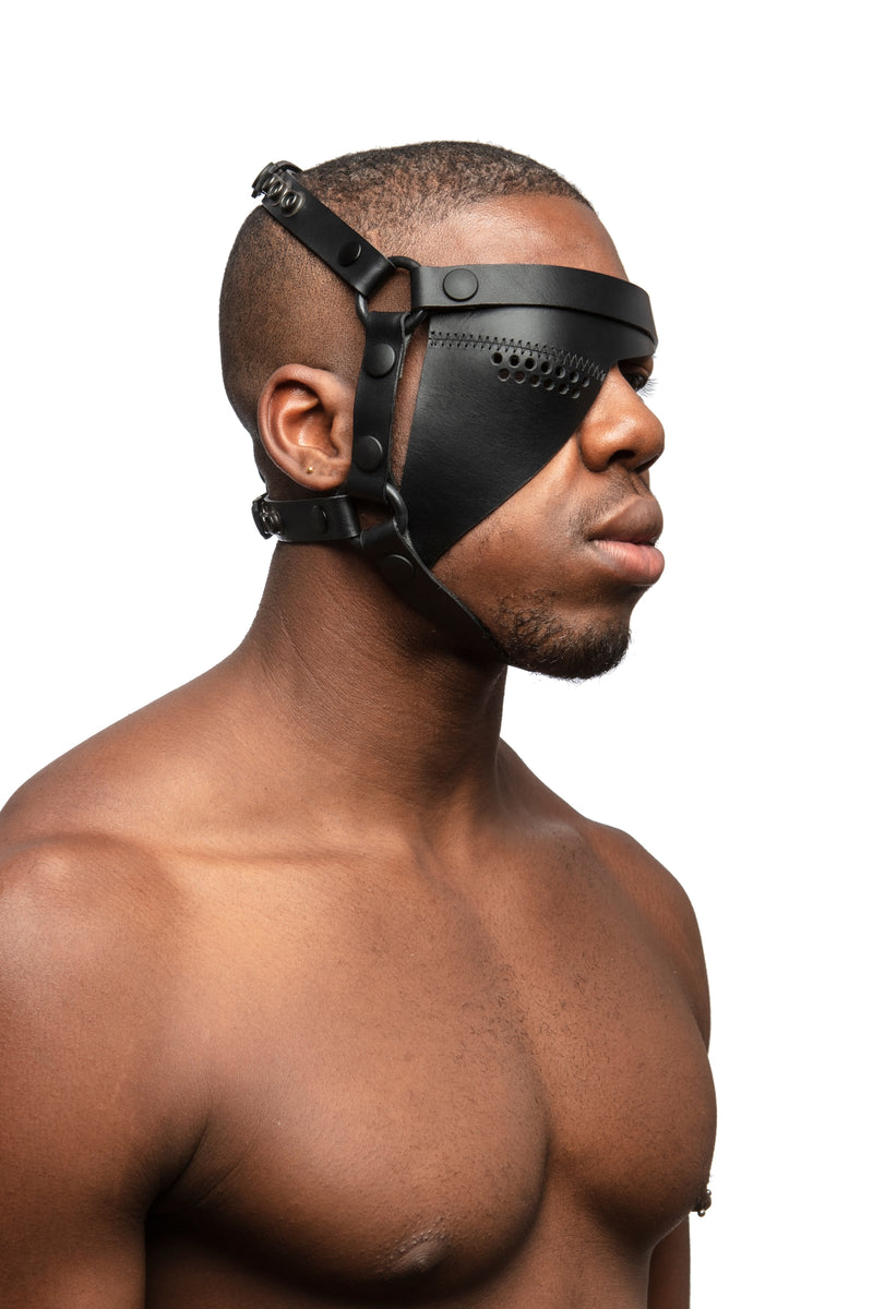 Model wearing black leather head harness and right eye patch with black metal hardware. Three quarter view.