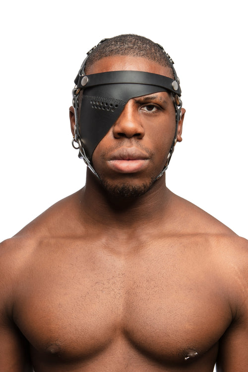Model wearing black leather head harness and eye patch with stainless steel hardware. Front view.